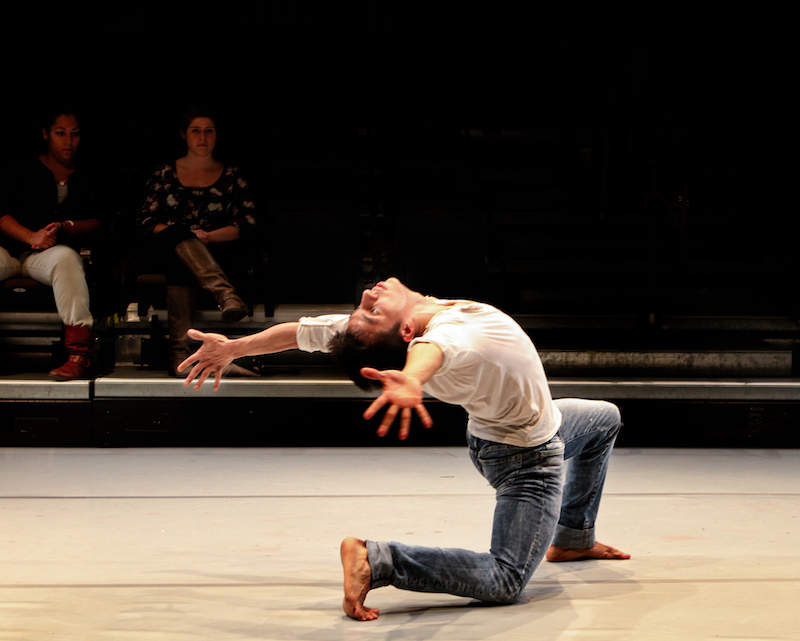 Victor Gonzalez in jeans and a long sleeve shirt kneels on one knee while in a dramatic back bend